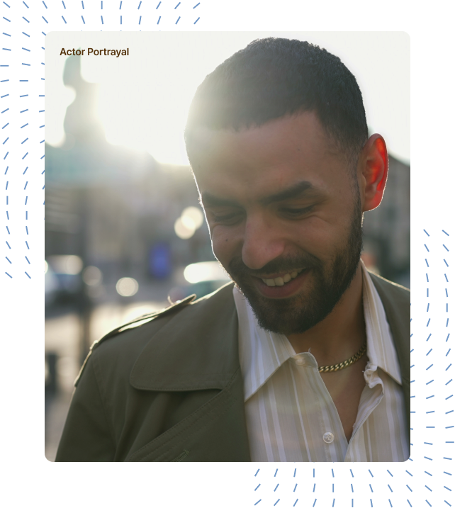 A man wearing an olive jacket standing outdoors, smiling with his gaze directed downward, while sunlight filters through the background, casting a warm glow around him.