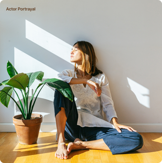 Woman in contemplative pose by a window with sunlight casting shadows, near a lush potted plant.